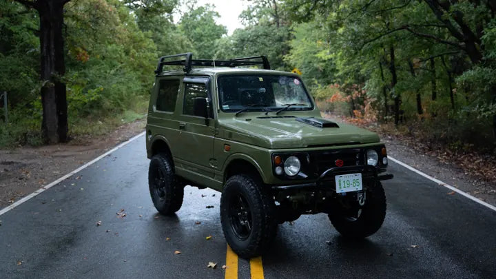 Front three-quarter view of the green Suzuki Jimny on a forest road